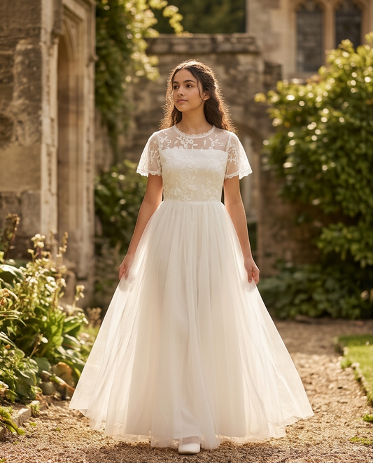 A girl stands on a sunlit garden path wearing a white lace midi dress with ruffled cap sleeves, a high neckline, and a full skirt. 