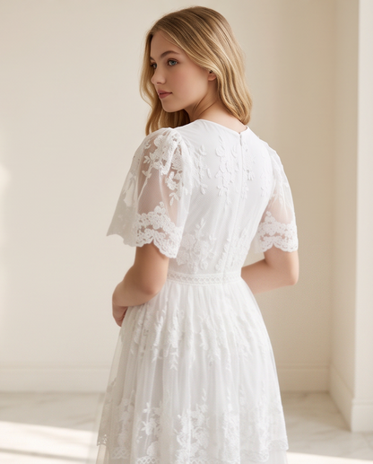 A woman wearing a white lace dress, photographed from the back in a softly lit indoor setting with cream walls and light-colored flooring.