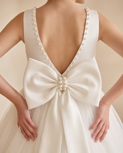Close-up back view of a white bridal gown with a deep V back, pearl bead trim, and a large satin bow at the center adorned with pearls, worn by a model.