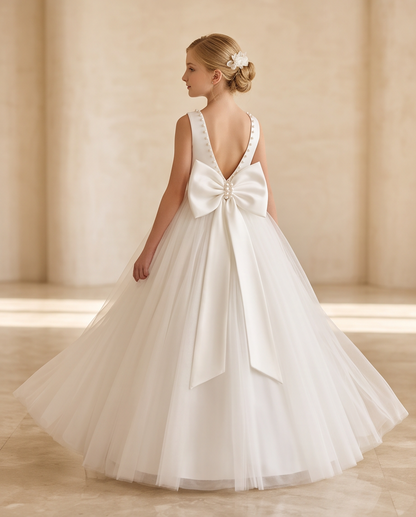 A young girl is shown from the back wearing a white sleeveless ball gown with a large bow at the back, photographed in a warm beige indoor setting with soft sunlight and columns.
