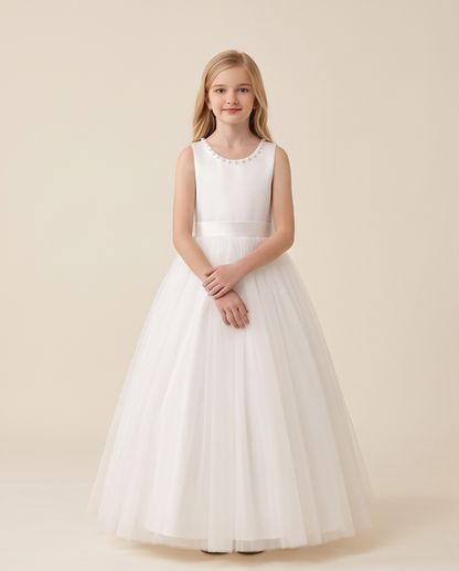 A young girl stands against a warm beige studio background wearing a sleeveless white formal dress with a scalloped neckline, a fitted waistband, and a full tulle skirt.