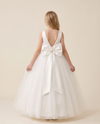 A young girl viewed from behind wearing a sleeveless white ball gown with a large satin bow at the back, photographed against a plain warm beige studio background.