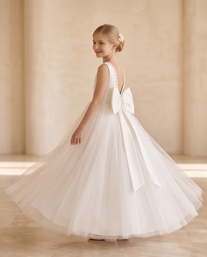 A young girl wearing a white sleeveless ball gown with a large bow on the back, photographed in a softly lit indoor setting with warm beige walls and columns.