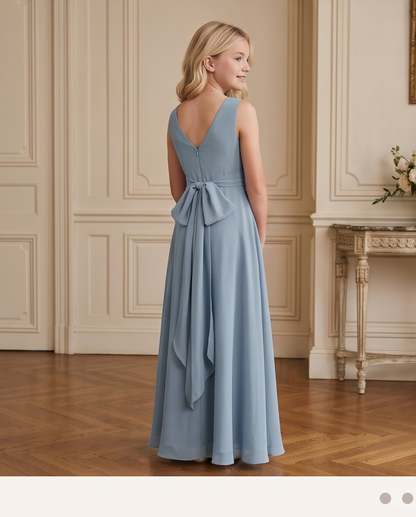 A young blonde woman stands in a classic indoor room, viewed from the back, wearing a floor-length dusty blue dress with a large bow detail at the back waist.