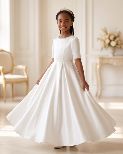 A young girl wearing a white first communion dress with short sleeves and a pleated skirt, standing in a bright, elegant room and twirling to show off the dress. She is wearing a small tiara.