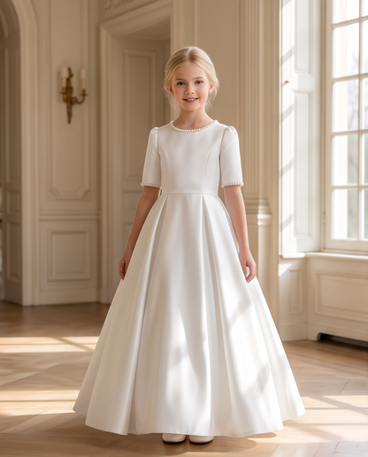 A young girl stands in a grand, sunlit interior wearing a floor-length white dress with short sleeves, a fitted bodice, and a full skirt. The room has cream walls, tall windows, and a light wood floor.