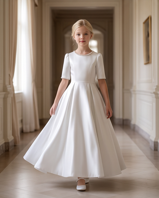A young girl stands in a grand, elegant hallway wearing a floor-length white satin dress with short sleeves, a round neckline, and a full skirt.