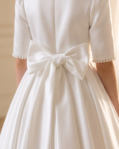 Close-up rear view of a white satin dress with short sleeves, pearl trim at the cuffs, and a large bow at the back waist.