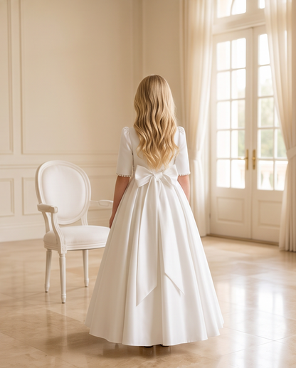A young girl with long blonde hair is seen from behind wearing a white short-sleeve ball gown with a large bow at the back, standing in a bright, elegant room with cream walls, a white chair, and French doors.