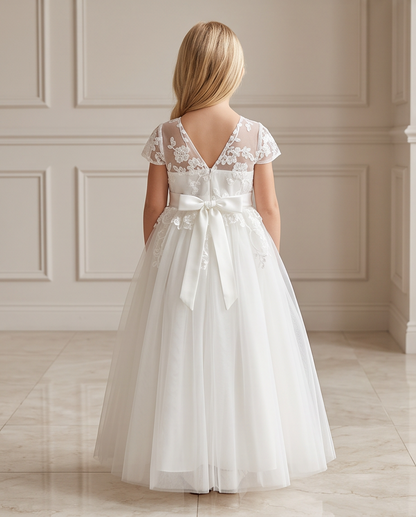 A young girl viewed from behind wearing a white lace and tulle formal dress with a large satin bow at the back, standing in an elegant indoor room with paneled walls and a tiled floor.