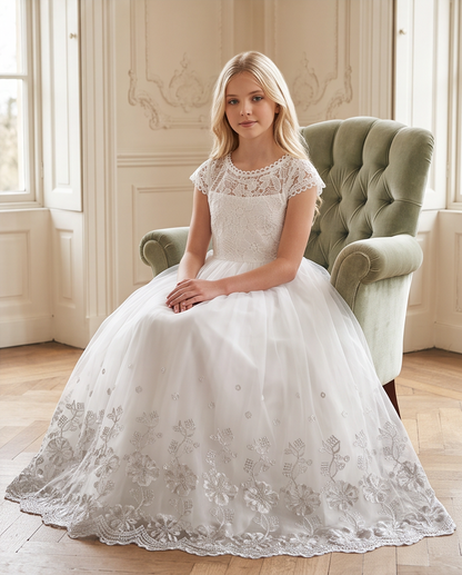 A young girl with blonde hair sits in a green tufted armchair wearing a formal white first communion dress with a lace bodice, cap sleeves, and a full embroidered tulle skirt in a bright room with wood floors and windows.