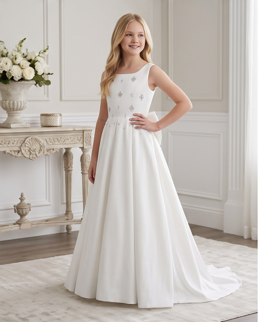 A young girl models a sleeveless white A-line dress with a fitted bodice, decorated with small silver floral appliqués, in an elegant, brightly lit room with a console table and white flowers.