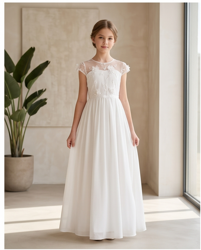 A young girl stands indoors wearing a long white formal dress with sheer cap sleeves, floral lace applique across the bodice, and a full flowing skirt. The room has warm beige walls, a large potted plant, and a window with bright sunlight streaming in.