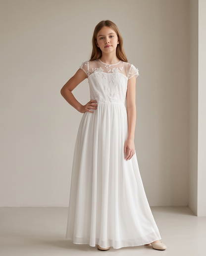 A young girl modeling a long white formal dress with a sheer lace bodice, cap sleeves, and a flowing chiffon skirt, standing in a minimalist beige room.