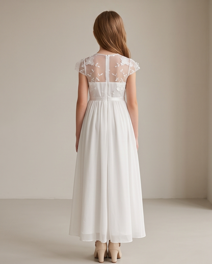A woman viewed from the back wearing a white A-line dress with a sheer floral lace back panel and cap sleeves, standing in a neutral studio setting.