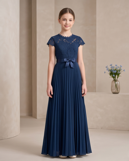 A young girl stands in a studio wearing a navy blue formal dress with a lace bodice, cap sleeves, and a pleated skirt with a matching waist bow. The background features warm beige columns and a small vase of blue flowers.