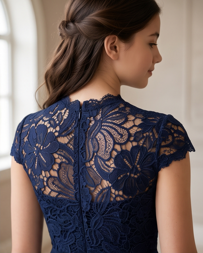 A woman shown from the back wearing a navy blue lace dress with floral lace patterns and a high lace neckline, photographed in a softly lit indoor setting.
