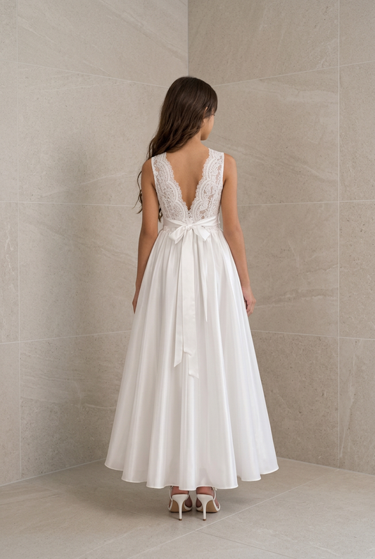 A woman viewed from the back wearing a sleeveless white maxi dress with a lace V-neckline, fitted waist, and a large bow at the back, standing in a tiled indoor setting.