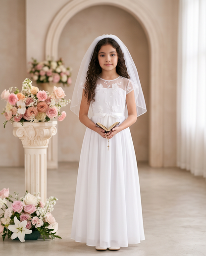 A young girl stands in a softly lit indoor chapel-like setting wearing a white first communion dress with a sheer veil. She holds a small book and rosary, with pink and cream floral arrangements and a decorative column beside her.