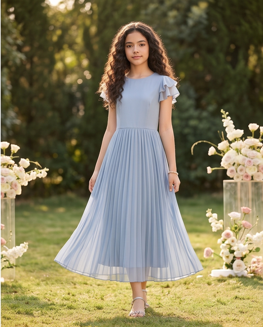 A young woman stands outdoors on grass wearing a light blue midi dress with ruffled cap sleeves and a flowing pleated skirt. The background shows blurred green foliage and flower arrangements in clear pedestals.