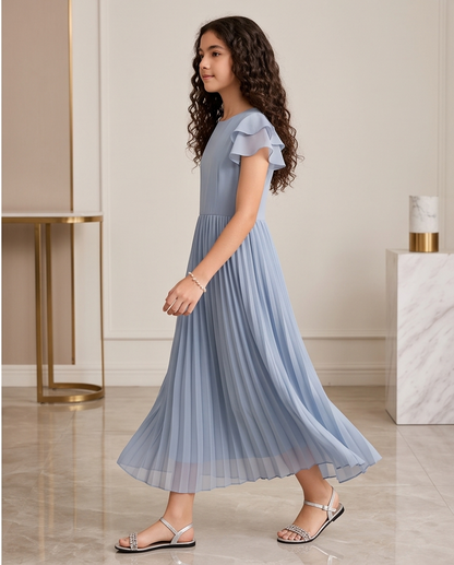 A young girl with long curly brown hair is wearing a light blue pleated dress with ruffled short sleeves and silver sandals, standing in a modern indoor room with marble floors and minimalist columns.