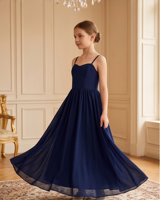A young girl wearing a navy blue sleeveless maxi dress with spaghetti straps and a sweetheart neckline, standing in a grand room with cream paneled walls and a chandelier.