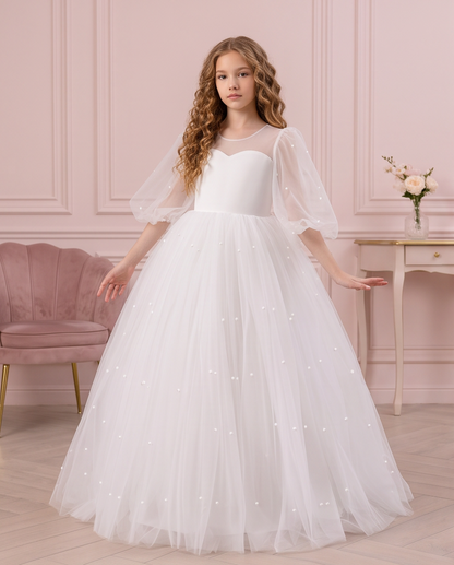 Girl wearing the Pearl Reverie white communion dress beside a floral fireplace, showing the full skirt.