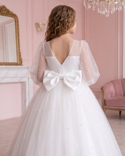 Girl wearing the Pearl Reverie white communion dress beside a floral fireplace, showing the full skirt.