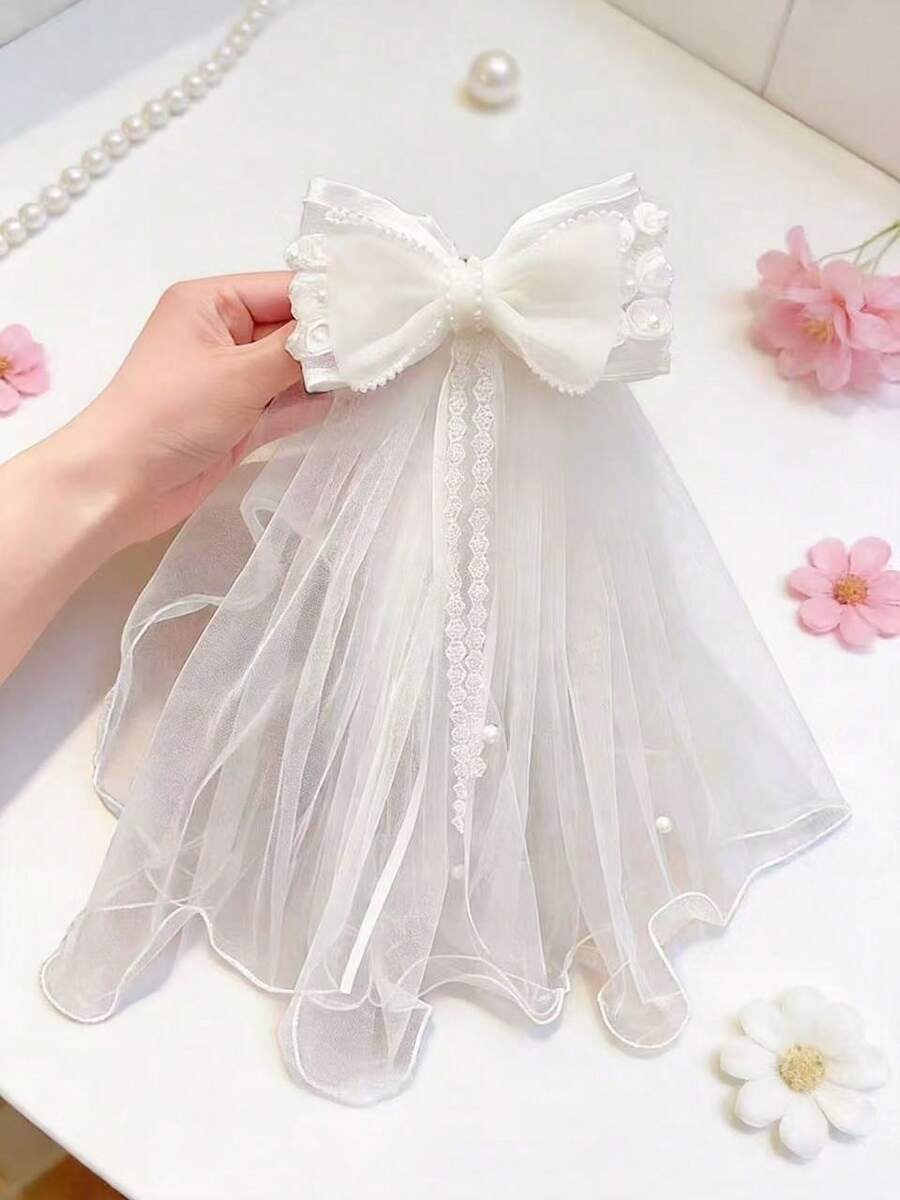 White ruffle organza bow communion veil with lace ribbon and pearl trim, flat lay on white surface with pink flowers