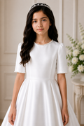 Young girl in a white dress with a tiara, standing indoors with a neutral background