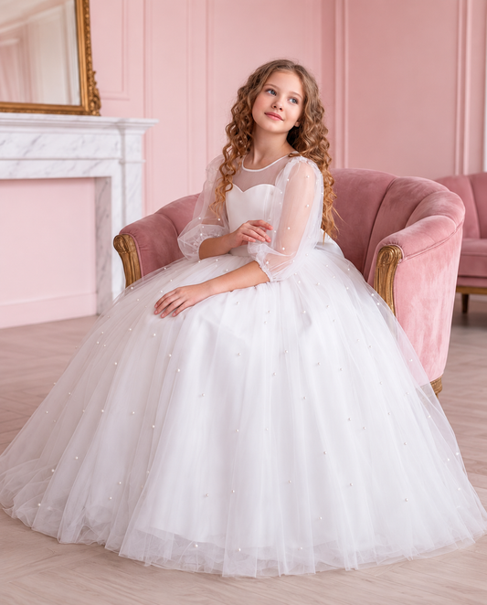 Girl seated in the Pearl Reverie white communion dress with sheer puff sleeves and a pearl-detailed tulle skirt.