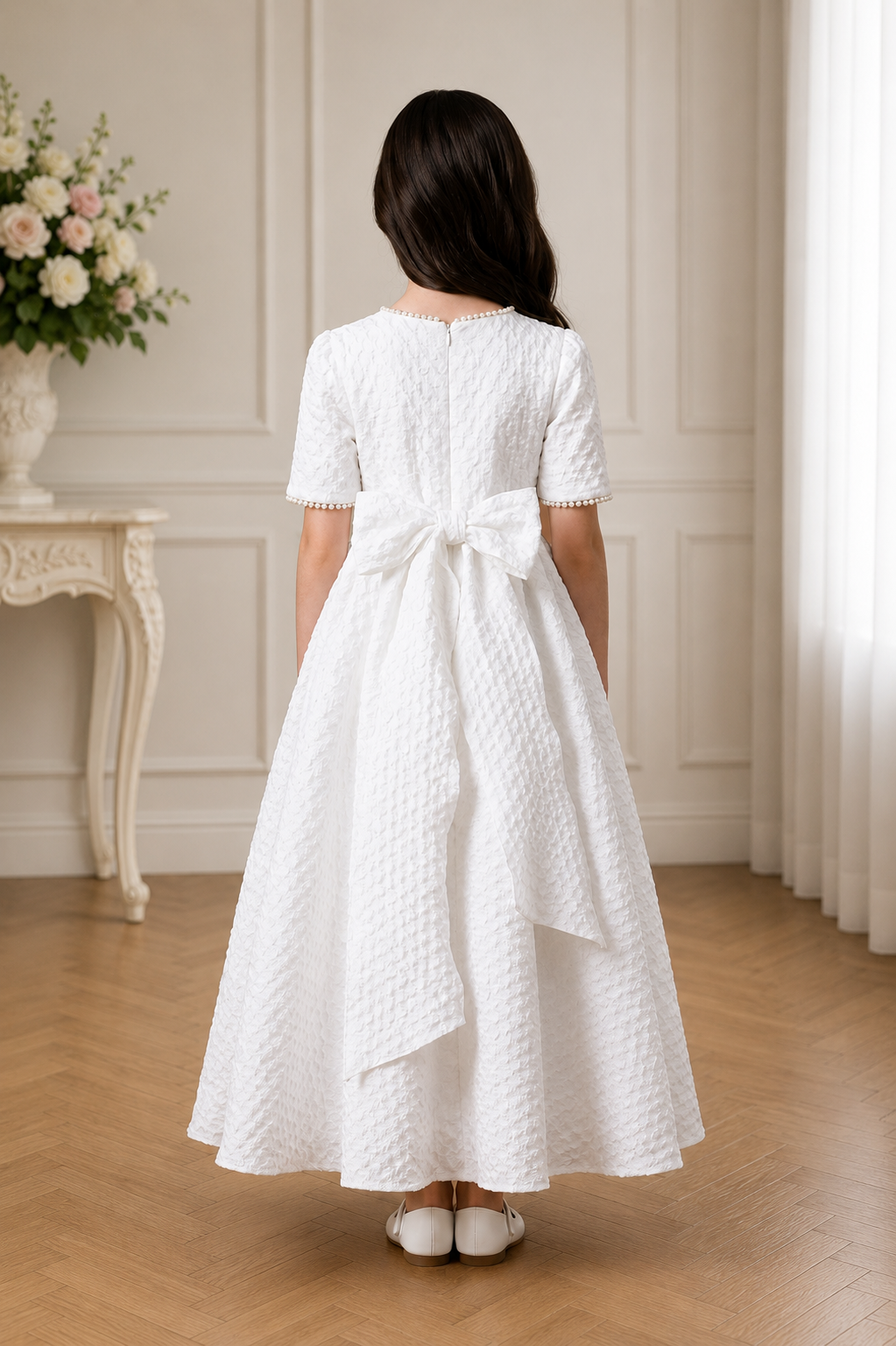 Girl wearing a white dress with a bow in a room with floral decorations.