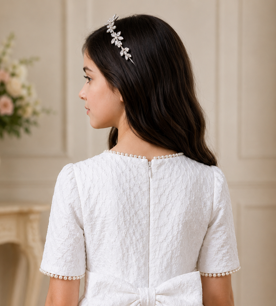Young girl wearing a white dress with pearl details and a decorative headband, standing against a neutral background.