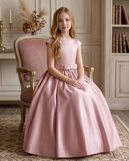 Young girl in a pink dress sitting on a chair in a room with books and decorative items.