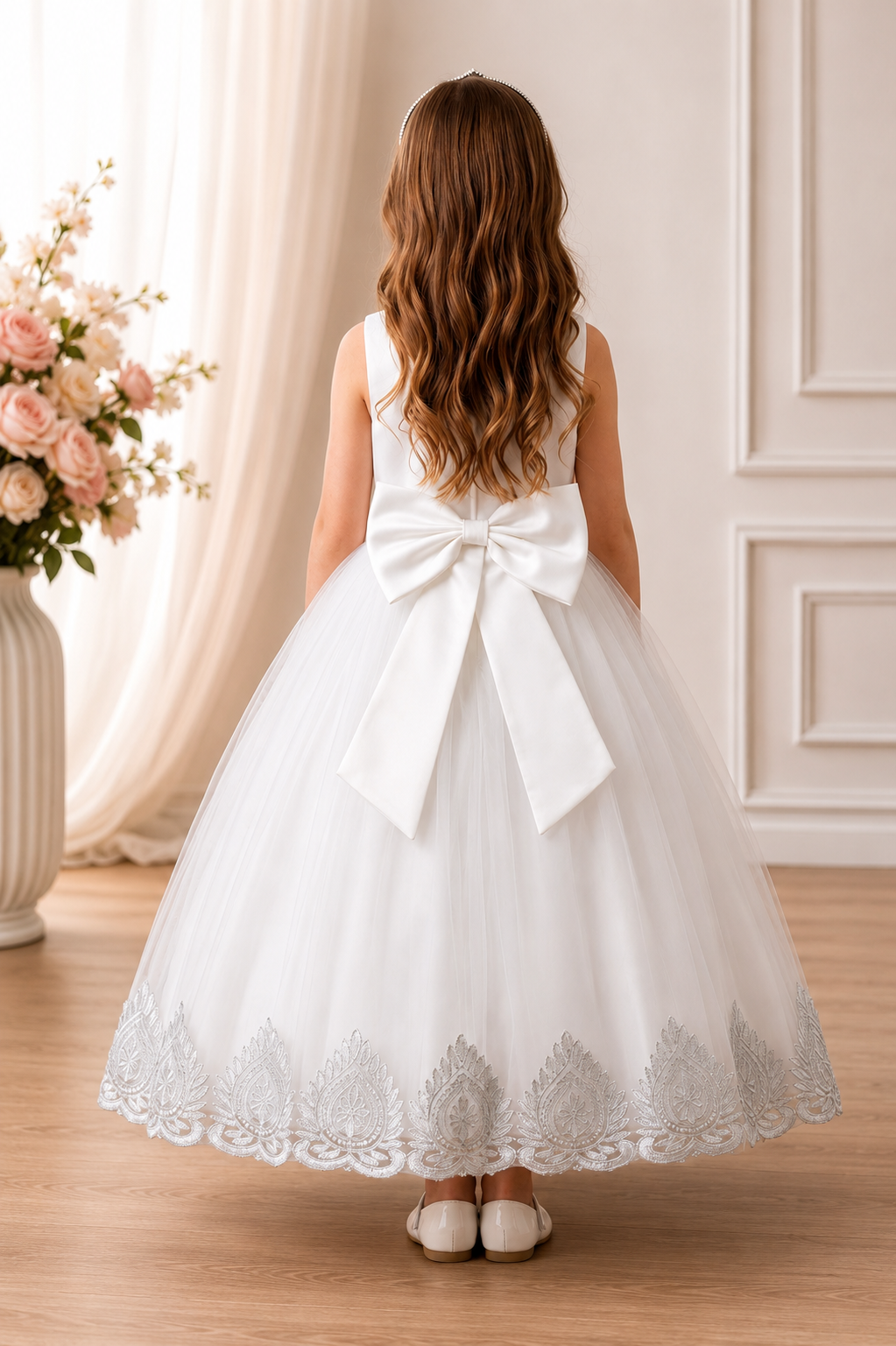 Young girl in a white lace dress with a large bow standing in a room with floral decorations.