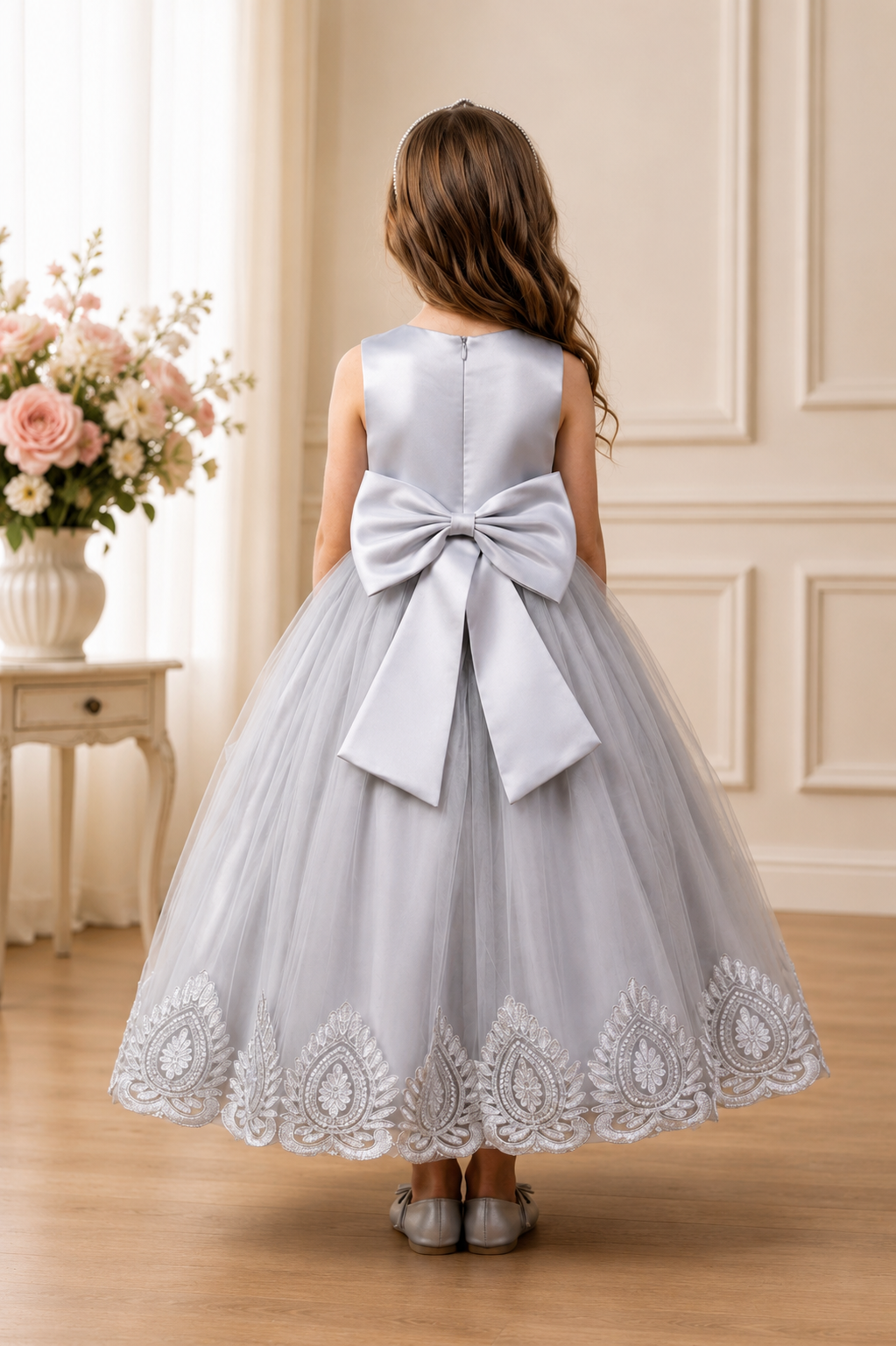 Young girl in a silver dress with a large bow standing in a room with floral decorations.