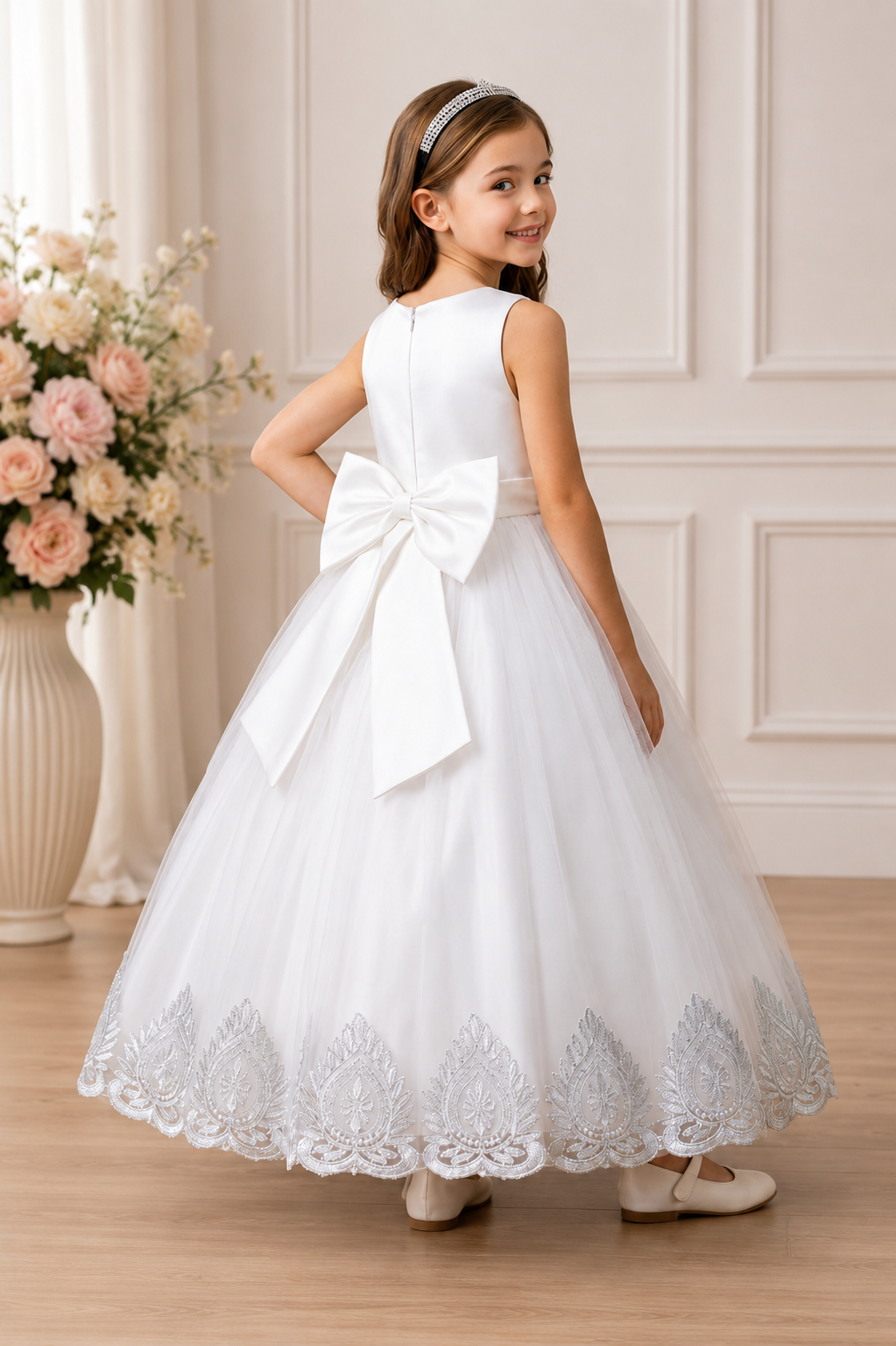 Young girl in a white lace dress with a large bow standing in a room with floral decorations.