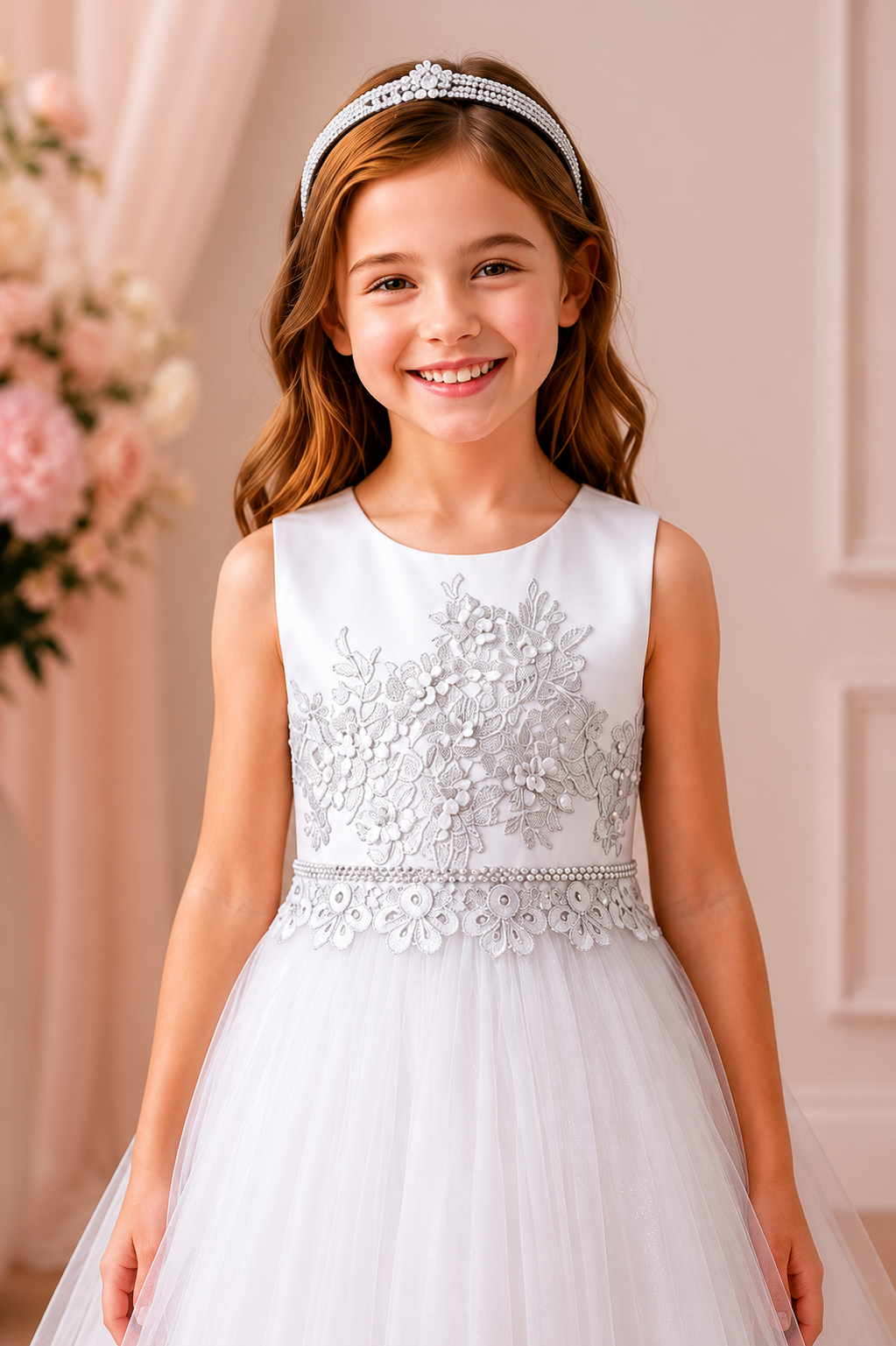 Young girl in a white floral dress with a tiara, standing against a soft pink background.