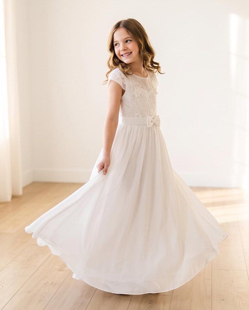 A young girl with wavy brown hair wearing a white lace and tulle first communion dress, captured mid-twirl in a bright, sunlit room with light wood floors.