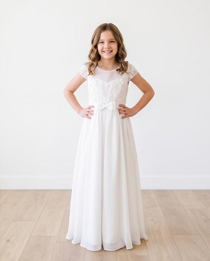A young girl stands with hands on hips wearing a long white lace and chiffon formal dress in a bright room with light wood floors and a white wall.