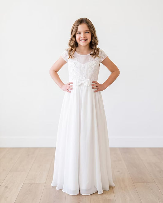 A young girl stands with hands on hips wearing a long white lace and chiffon formal dress in a bright room with light wood floors and a white wall.