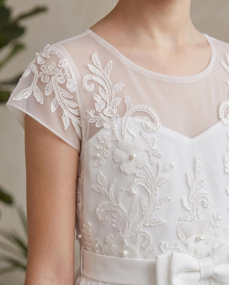 Close-up of a white dress with sheer mesh cap sleeves, intricate white floral lace embroidery, and pearl-like bead accents.