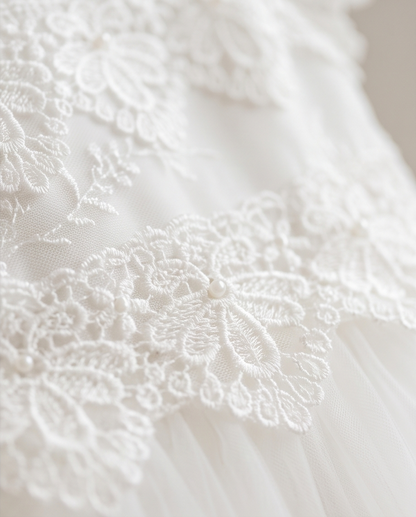 Close-up of a white lace and tulle garment detail, showing floral lace appliqué over a soft tulle base with small pearl-like accents.