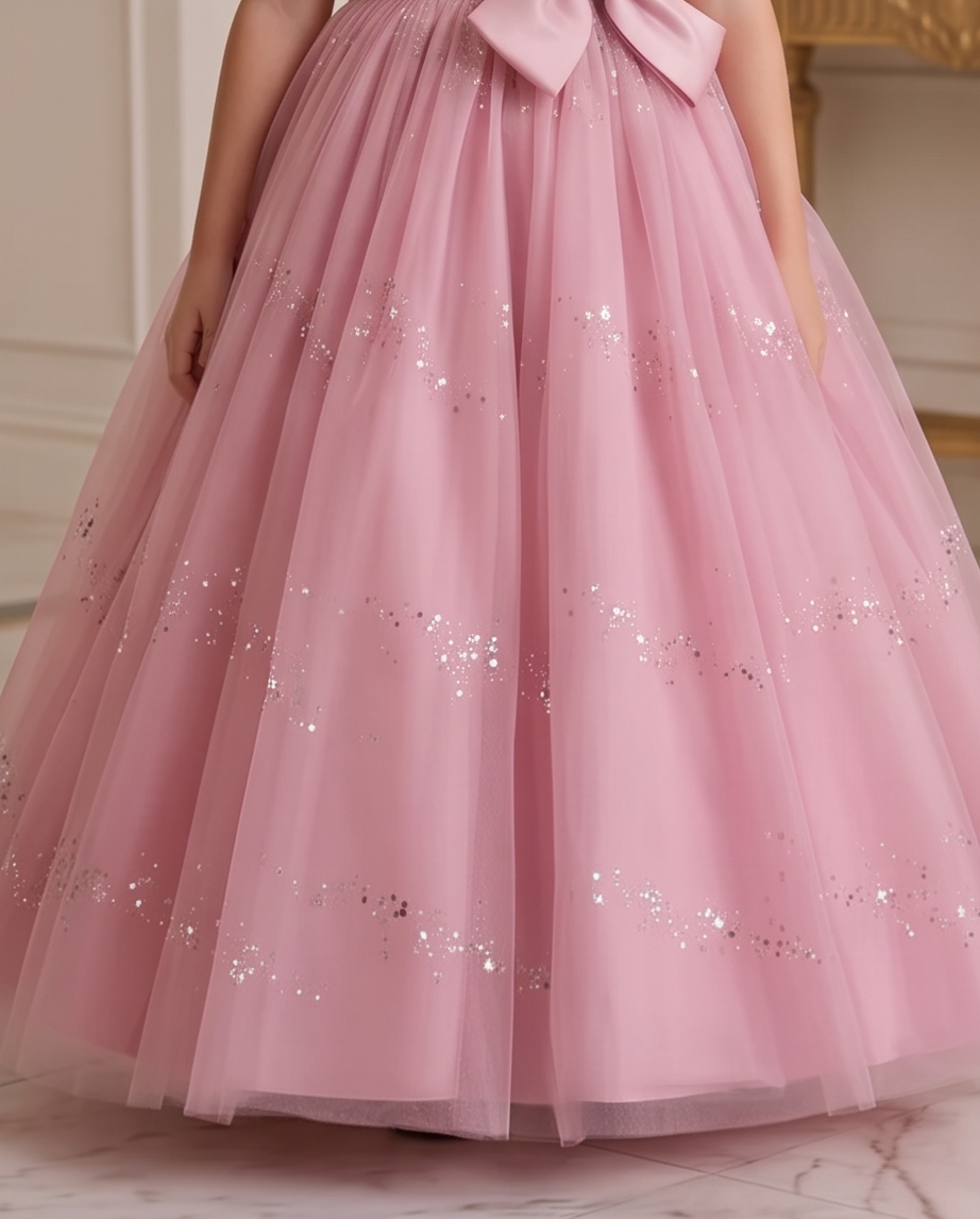 A pink tulle ball gown with a large bow at the waist and scattered silver sequins, shown on a child from the waist down in an indoor setting.
