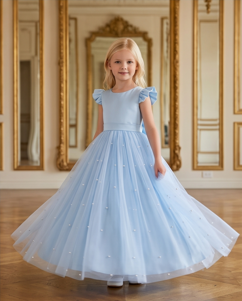 A young girl with blonde hair stands in a grand room with gold-framed mirrors, wearing a light blue ballgown with ruffled cap sleeves and a full tulle skirt decorated with small pearl-like accents.