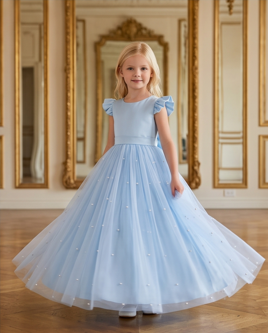 A young girl with blonde hair stands in a grand room with gold-framed mirrors, wearing a light blue ballgown with ruffled cap sleeves and a full tulle skirt decorated with small pearl-like accents.