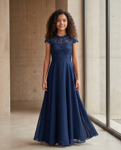 A young girl with curly brown hair stands in a modern concrete-walled room wearing a navy blue formal gown with a lace bodice and flowing chiffon skirt.