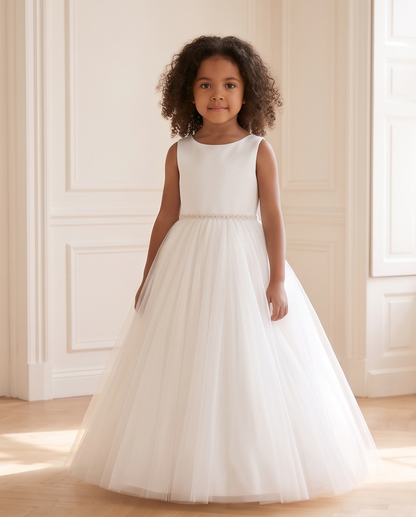 A young girl with curly dark hair stands indoors wearing a sleeveless white formal dress with a fitted bodice, a thin embellished waistband, and a full tulle skirt.