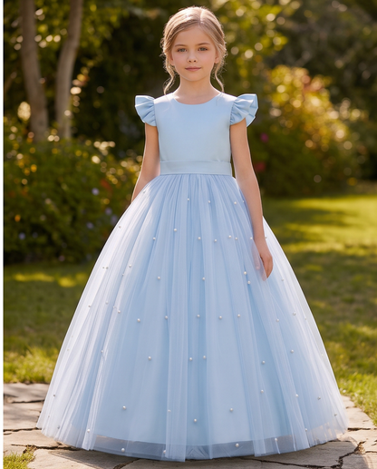 A young girl stands outdoors in a lush garden wearing a light blue ball gown with puffed cap sleeves, a fitted bodice, and a voluminous tulle skirt adorned with small pearl-like embellishments.