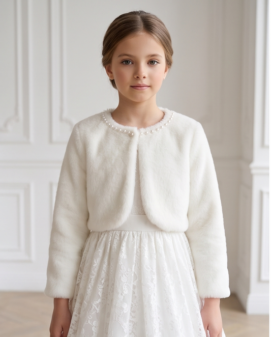 A young girl with light brown hair is wearing a white faux fur cropped jacket with pearl trim and a white lace dress, standing in front of a white paneled wall.